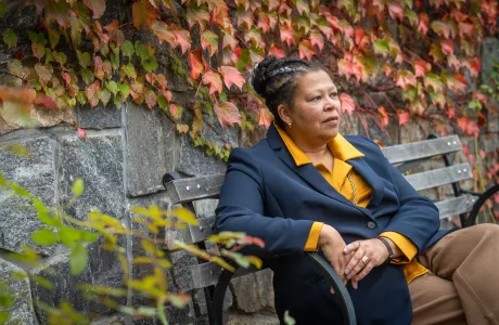 Photo of Sarah Willie-LeBreton sitting on a bench and looking into the distance, with fall foilia