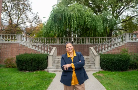 Photo of Sarah Willie-LeBreton smiling and standing outside with her arms folded, in front of the stone stairs in The Quad. 