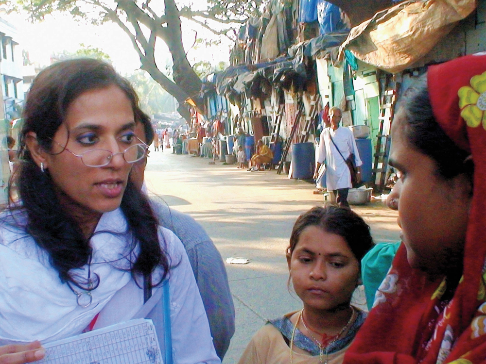 A woman with shoulder length black hair in a white sari speaks to a person in a red sari and a child