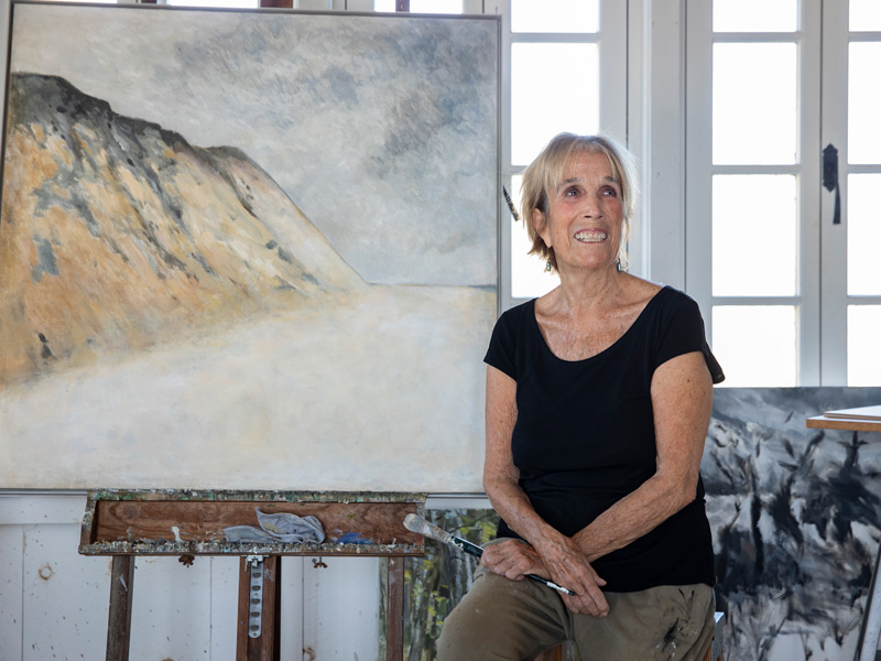 Anne Peretz holding a paint brush, smiling, in front of a tranquil painting of a sand dune