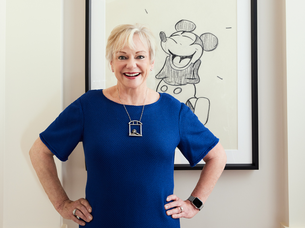 Christine McCarthy, smiling, with her hands on her hips, in front of a framed Mickey Mouse sketch