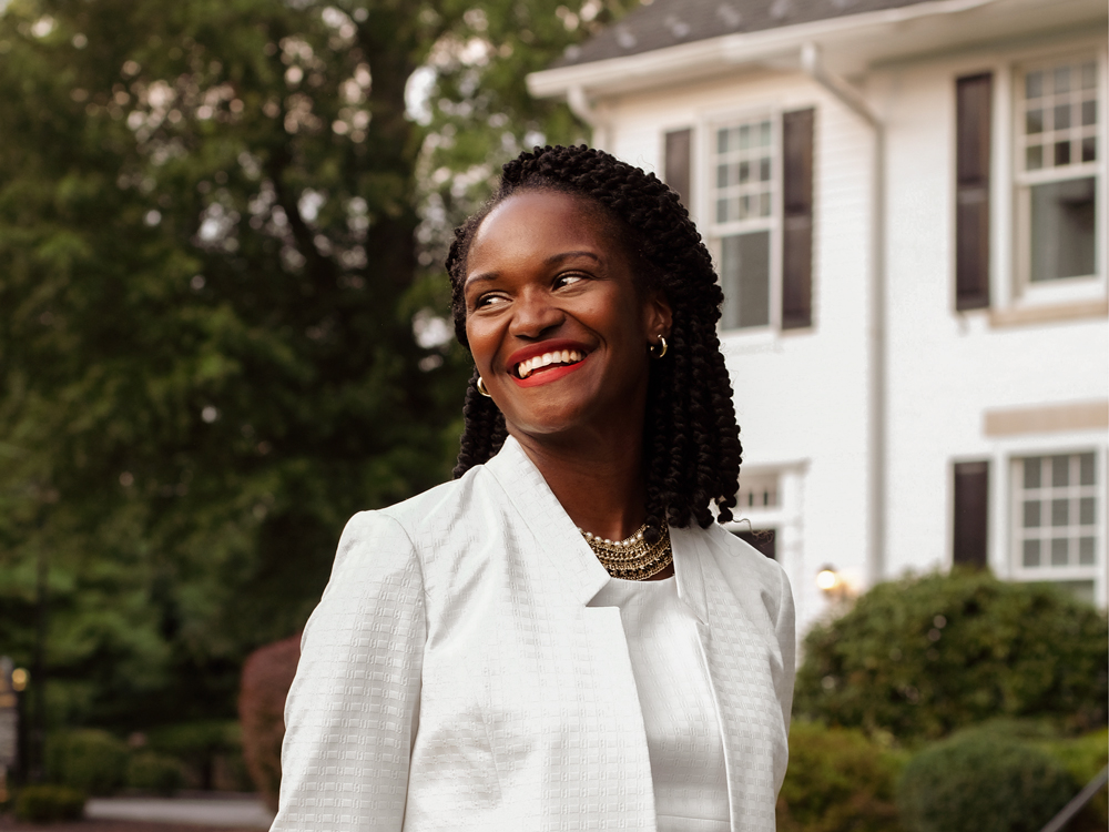 KaNeda Bullock in a white jacket and dress in front of a suburban colonial house
