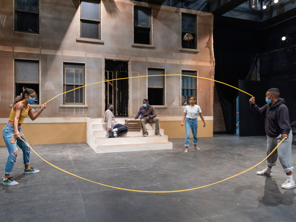 Actors turning double dutch ropes on a theatre set with a brownstone set in the background.