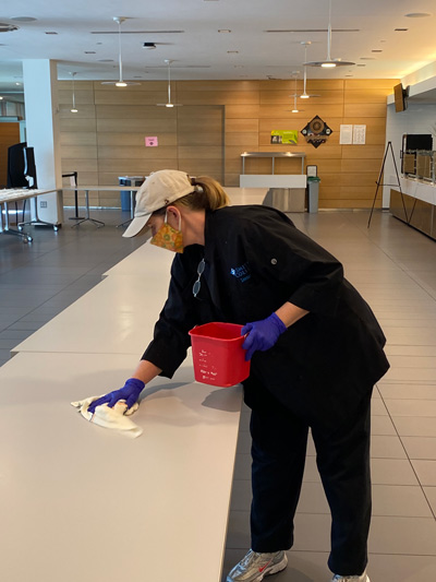 Masked, uniformed dining worker wiping down a table in the Campus Center