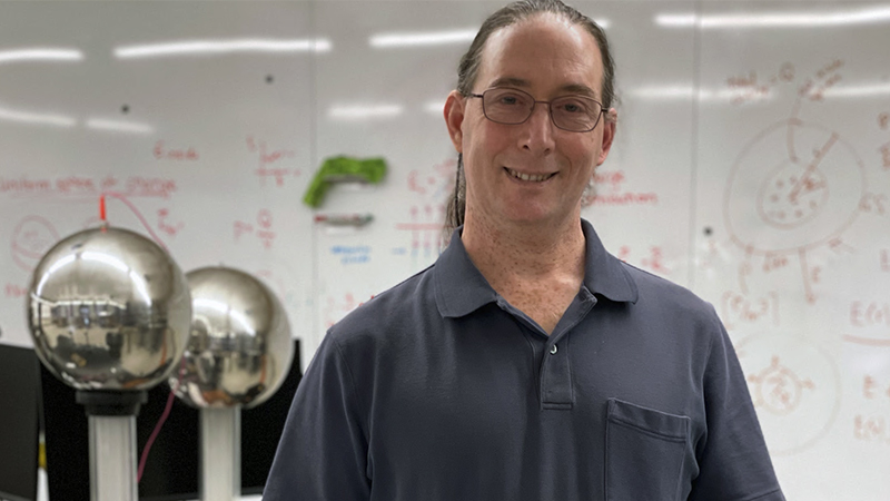 A middle aged white male professor with glasses in a dark gray-blue polo short-sleeved collared shirt stands in front of a white board in a college laboratory.