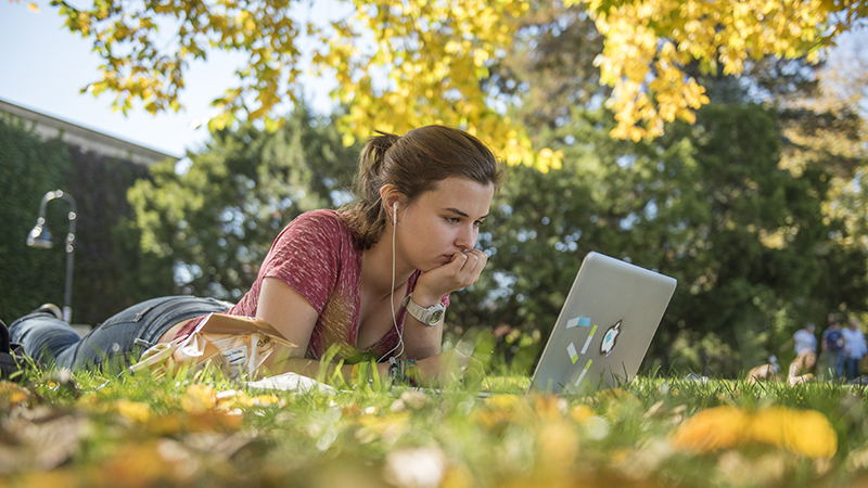 Student outside lying on the grass looking at a laptop