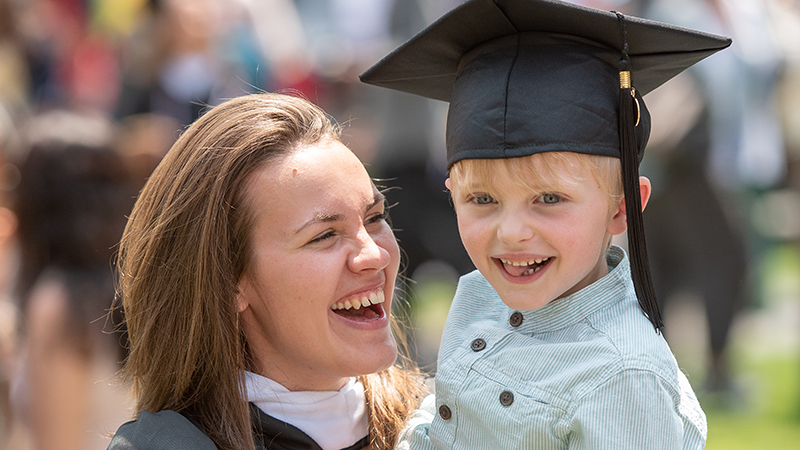 Graduating student in their regalia holds a young child wearing their graduation cap