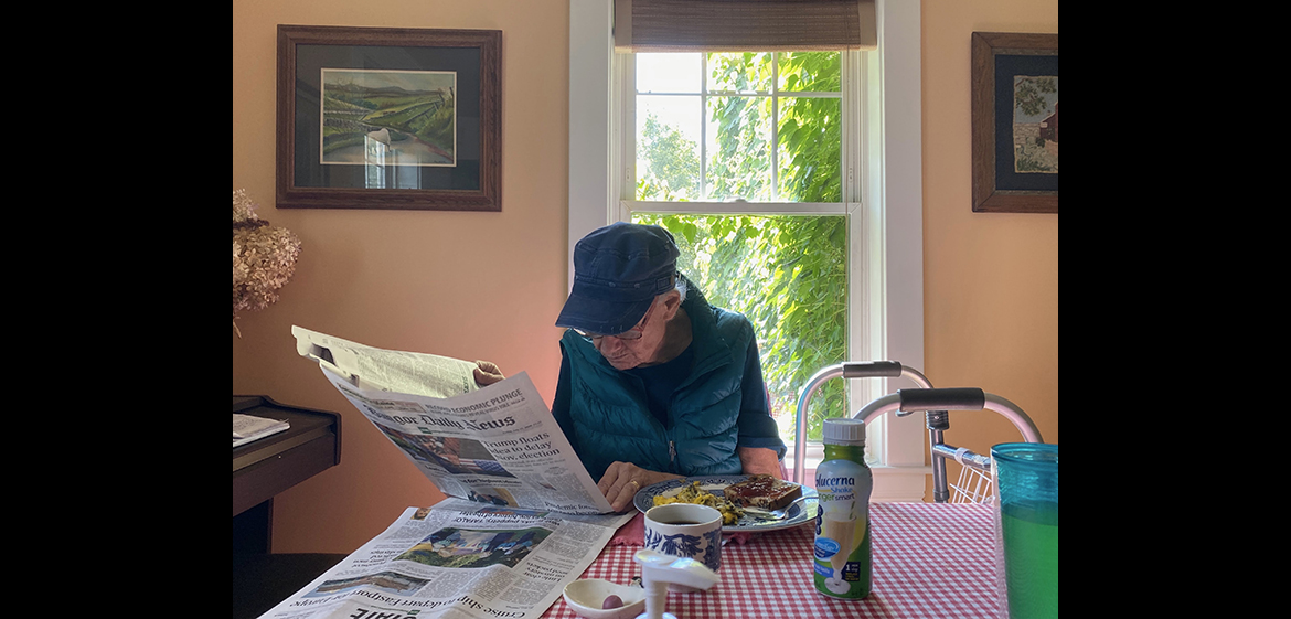 Photo of an older adult reading a the Bangor Daily News and eating a breakfast of scrambled eggs and toast
