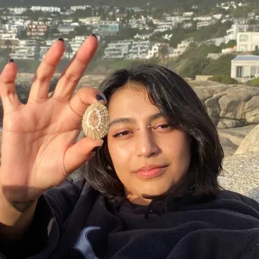 Tiki Pather holds up a shell and looks at the camera with white buildings visible behind
