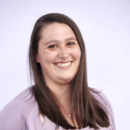 A woman with long brown hair smiles at the camera. She is wearing a light purple cardigan and blouse.
