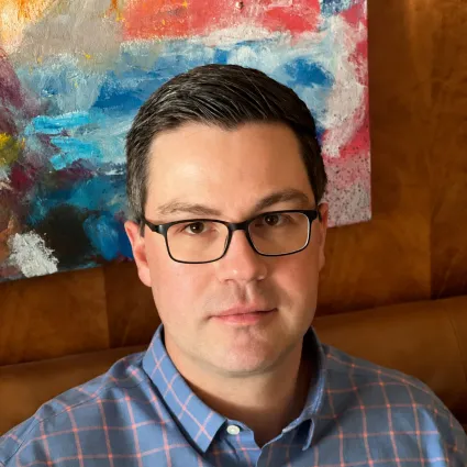 Portrait of a man with short dark hair, black-rimmed glasses, and a light blue and pink patterned button-up shirt, seated in front of a colorful abstract painting.