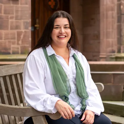 Portrait of a smiling woman with long dark hair, wearing a white button-up shirt with a green scarf draped around her neck, seated on a wooden bench outdoors in front of a stone building.