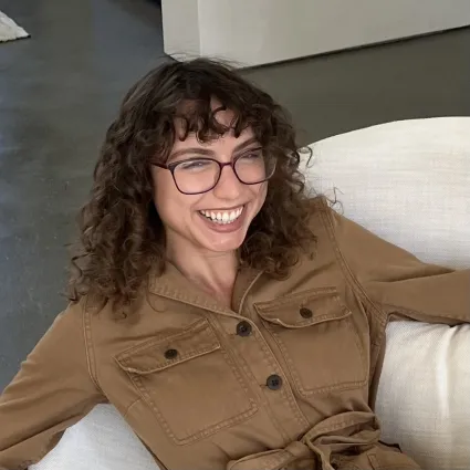 Headshot of a person with curly dark hair and glasses laughing while leaning back on a white sofa.