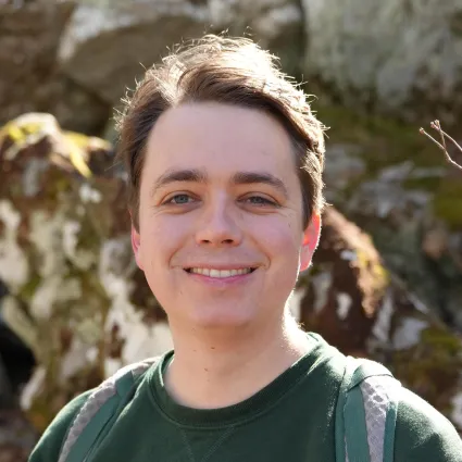 A close-up portrait of a person smiling directly at the camera while wearing a dark green long-sleeved shirt and backpack straps. Sunlight catches the back of their hair, with a softly blurred, rocky outdoor terrain in the background.