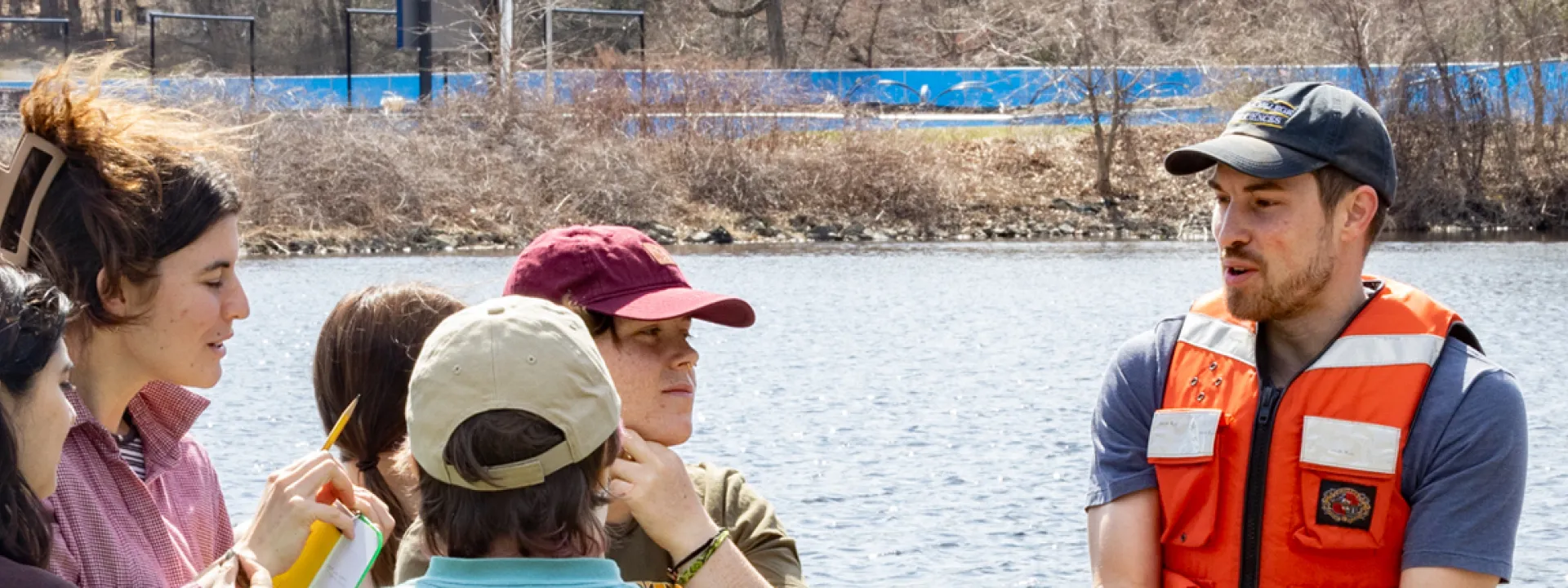 A wide, panorama-style photograph of a group of students and instructors gathered outdoors by the edge of a river or lake. On the right, a person wearing a blue baseball cap and a bright orange safety vest speaks to the group. On the left, several students listen closely, with one person writing in a notebook using a pencil. The background features calm, sunlit water backed by bare trees and campus athletic fields under a bright sky.