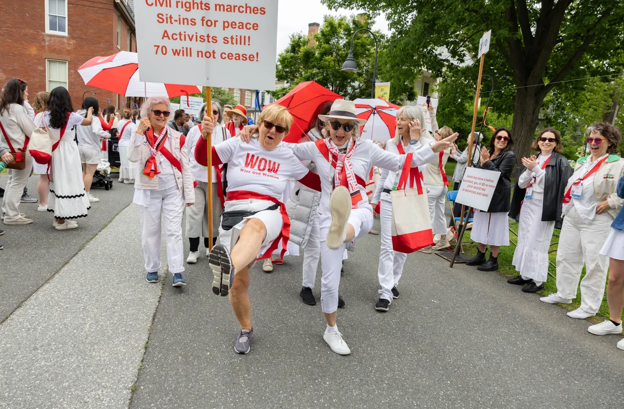 Alums from the class of 1970 wearing white and red march in the Reunion II parade. Two have their arms around one another's shoulders and they are enthusiastically kicking their legs.