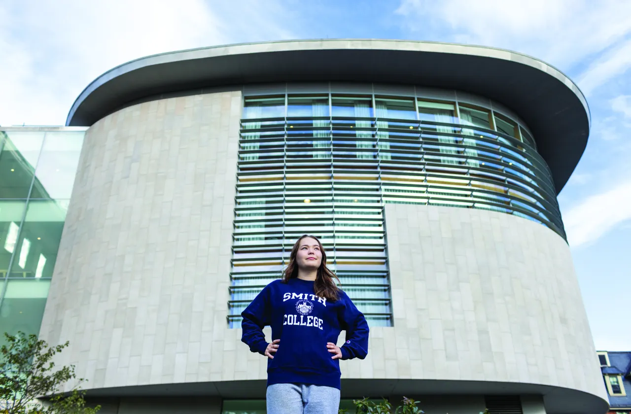 A student wearing a Smith College sweatshirt stands with her hands on her hips in front of one of the wings of Neilson Library