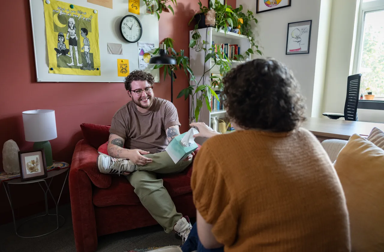 A counselor sits on a chair and hands a client a box of tissues