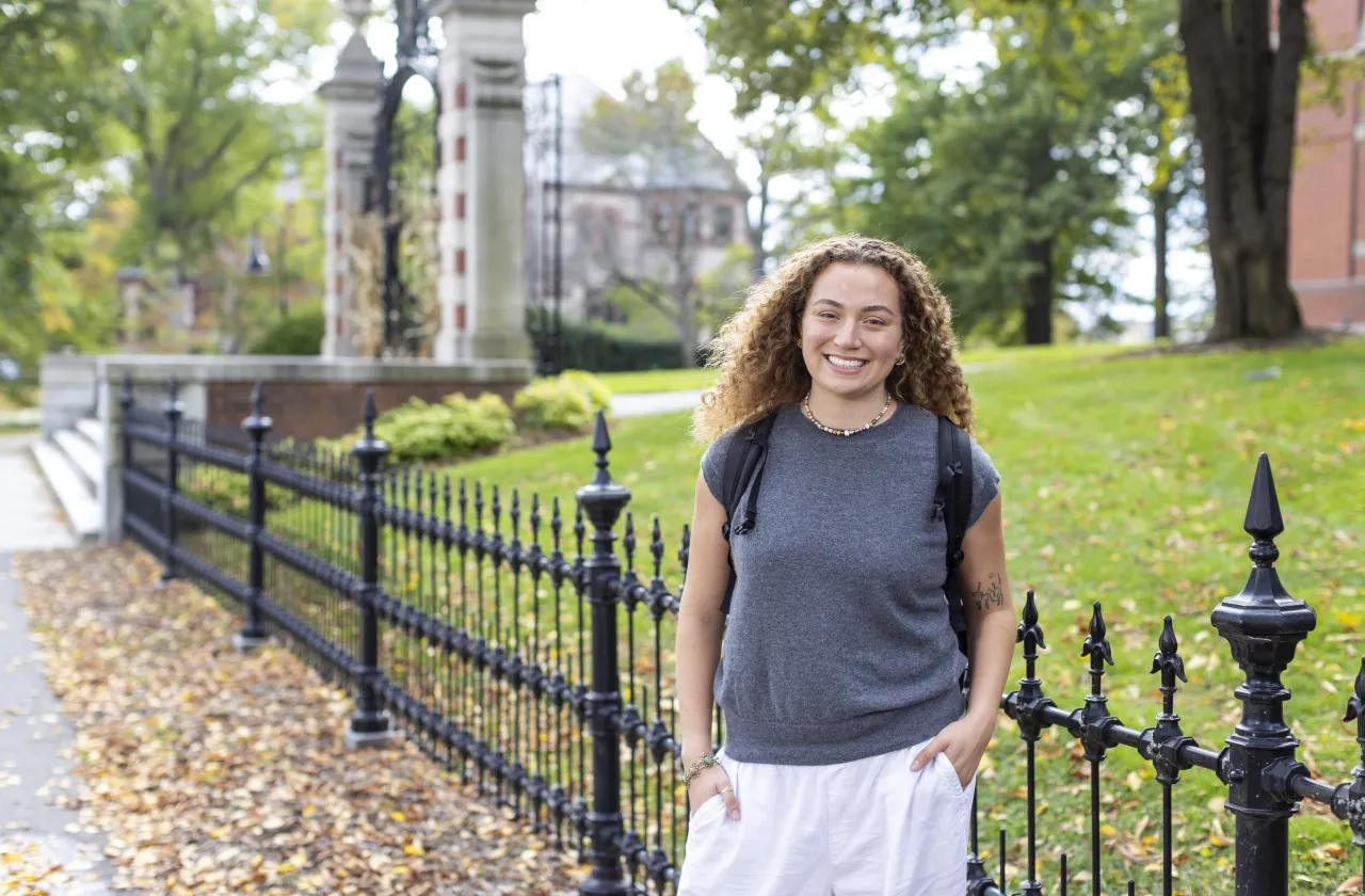 A smiling student with curly hair wearing a gray top and backpack stands outdoors along a black wrought-iron fence on a college campus.