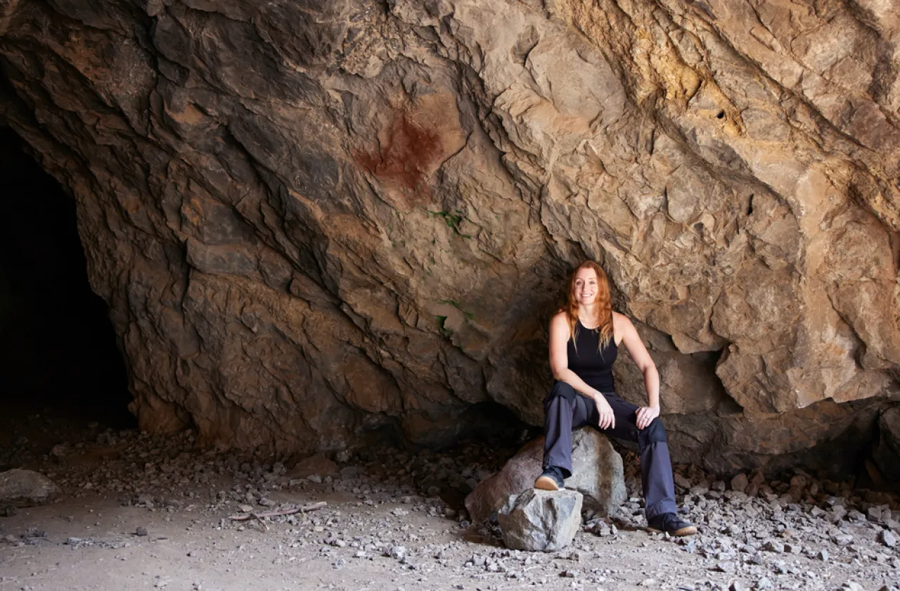 Jess_Phoenix, smiling, sitting on a rock at the mouth of a cave