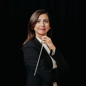 A woman in a black suit confidently holds a conductor's baton against a dark background. Her expression is focused and poised, conveying leadership.
