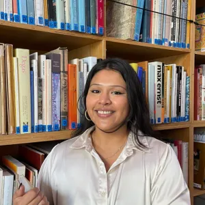 A young woman with dark hair smiles in front of a colorful bookshelf filled with books, wearing a light blouse and hoop earrings.