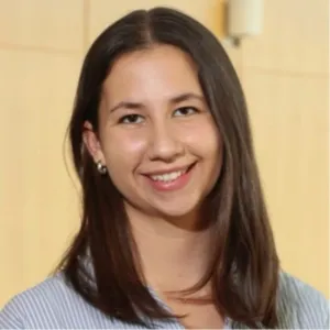 A headshot of a young woman with long, straight dark brown hair smiling at the camera. She is wearing a light blue and white striped button-down shirt and small silver hoop earrings, positioned against a neutral, warm wood-paneled background.