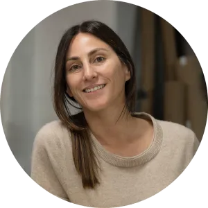 Portrait of a smiling woman with long brown hair parted down the middle and a beige knit sweater, set against an indoor background.