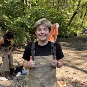 Danika Lipchitz smiling and giving two thumbs up while wearing Cabela's chest waders in a wooded riverbed.