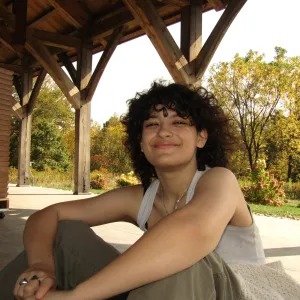 Ellie Paximadas sitting cross-legged under a wooden pavilion on a sunny autumn day.