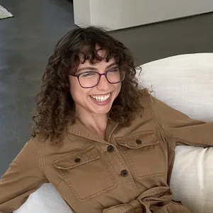 Headshot of a person with curly dark hair and glasses laughing while leaning back on a white sofa.