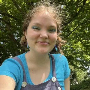 Outdoor selfie of Ronja Eilfort smiling softly, wearing blue eyeshadow, a bright turquoise shirt, and purple overalls under a canopy of green trees.