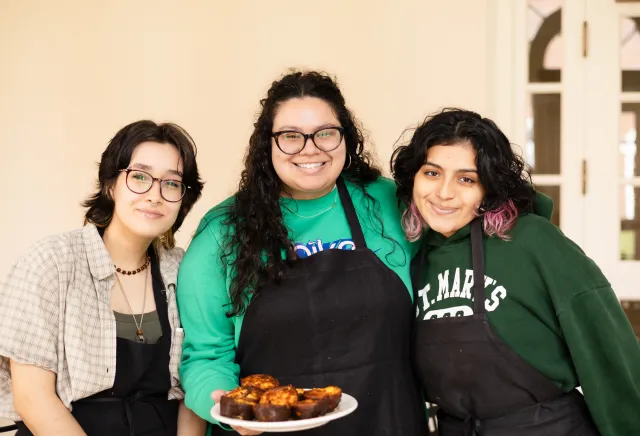 Three students in aprons smile while holding a plate of food on IS Day