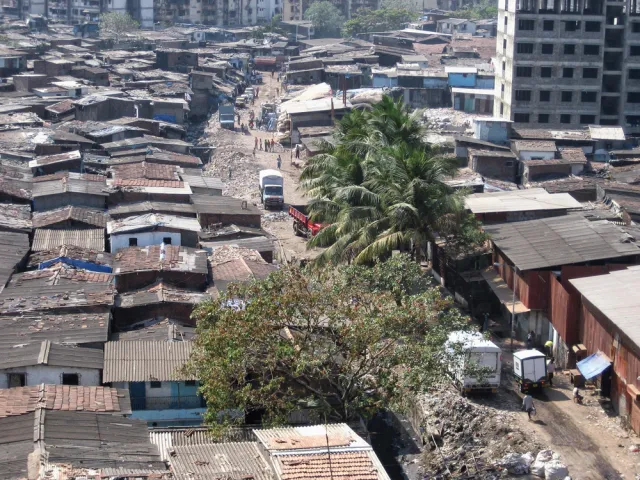Landscape color photo of concrete houses with tile roofs and a dirt road. There are palm trees, trucks and an tall empty building that could be abandoned or being constructed