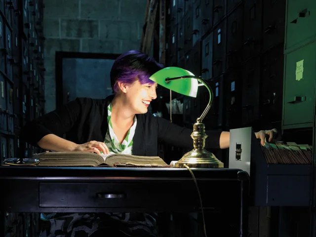Kory Behny Stamper looking in a filing cabinet while seated under a green table lamp.
