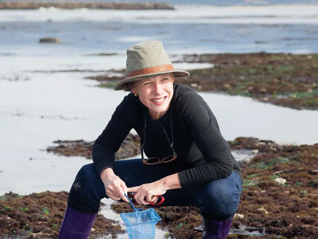 Mary Ellen Hannibal kneeling on coastal wetlands with a small net, smiling and squinting