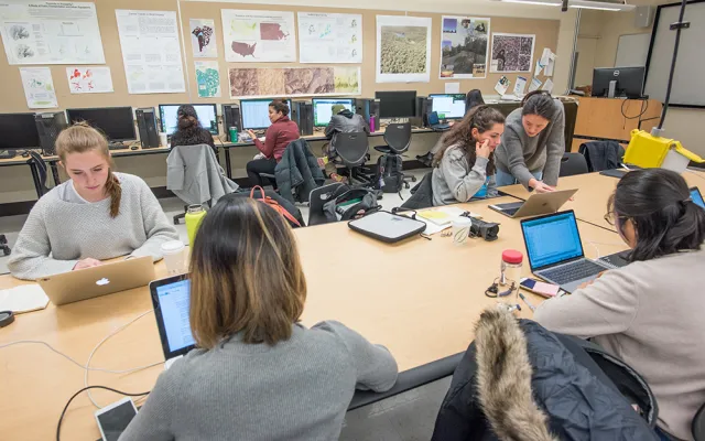 Multiple students work with laptops on a shared table while other students work on a row of desktop computers