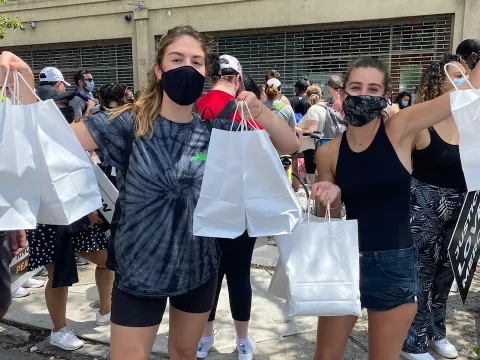 Two white friends wearing masks hold up bags of food