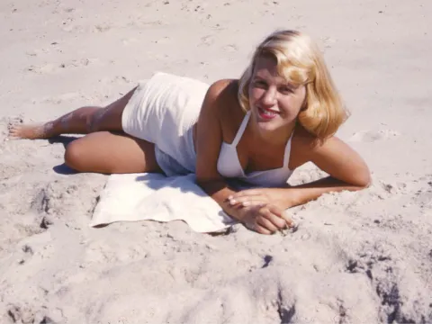 Sylvia Plath in a white bathing suit posing on the beach, smiling at the camera