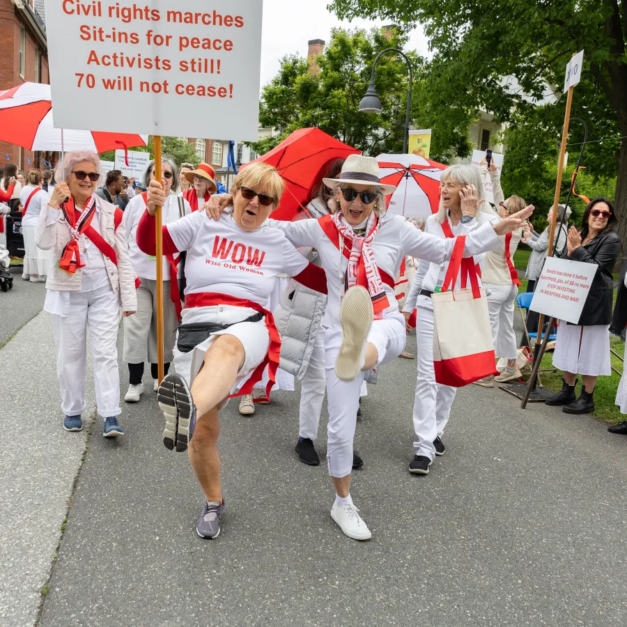 Alums from the class of 1970 wearing white and red march in the Reunion II parade. Two have their arms around one another's shoulders and they are enthusiastically kicking their legs.