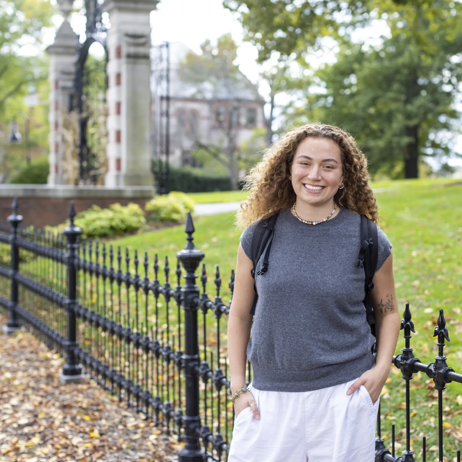 A smiling student with curly hair wearing a gray top and backpack stands outdoors along a black wrought-iron fence on a college campus.