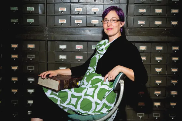 Kory Behny Stamper in a green dress with purple hair. She holds a large rare book in front of a bank of card catalogs