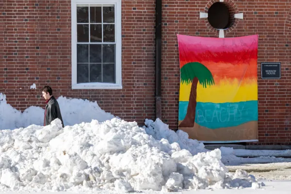 Person walking through snow drifts. Behind them is a brick building with a bright rainbow banner