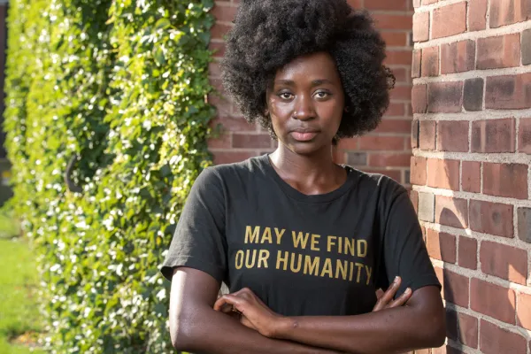 Hawa Tarawally leaning against a brick wall. Her shirt says, "May we find our humanity."