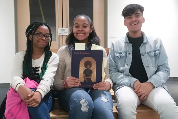 Three students of color seated and smiling at the camera. The middle person holds a copy of the exhibit book Black Refractions