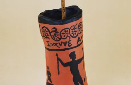 Tall ceramic vessel with an orange and black Greek-style design. Features human figures and patterns, topped with a numbered wooden rod.