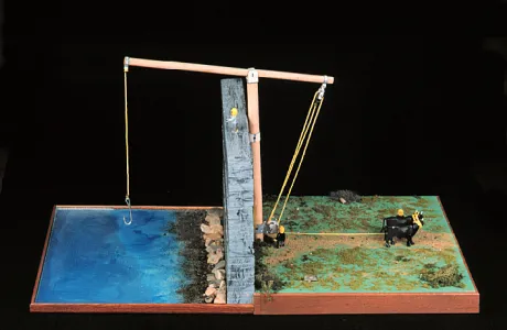 A wooden lever balances between a water-filled area on the left and a green field with an ox on the right