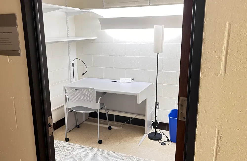 A small, well-lit lactation room with a white chair at a desk, shelves above, a floor lamp beside, a wall-mounted sign outside, and a blue bin on the floor.