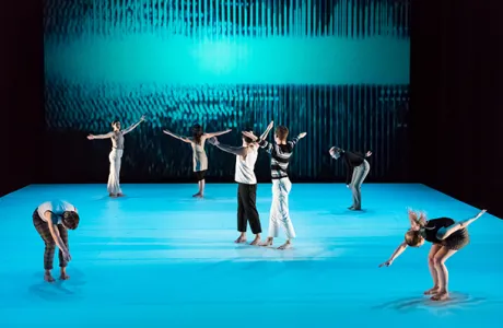 Seven dancers on stage against a bright blue floor and backdrop