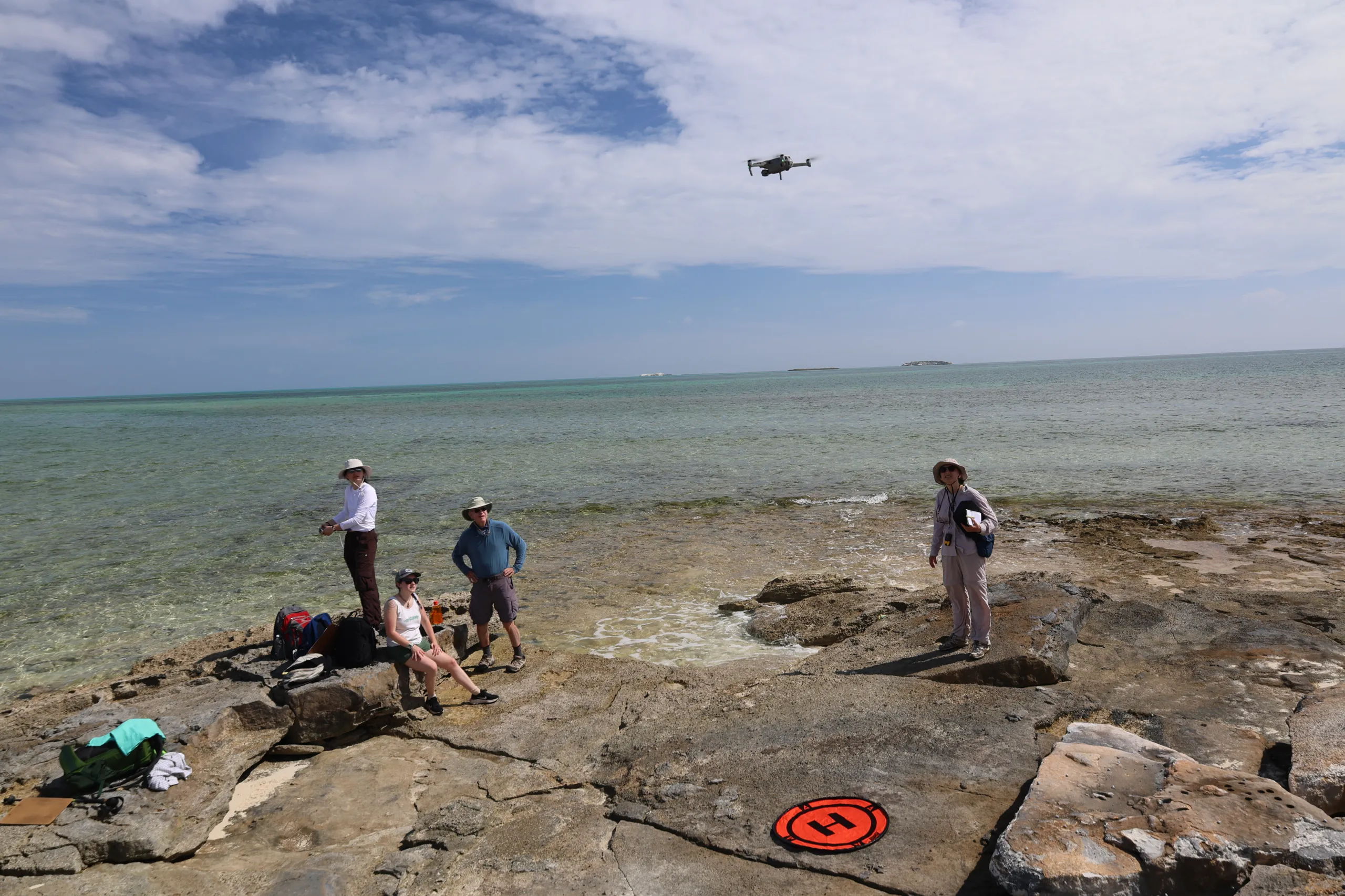 A group of four people stands on a rocky coastline beside clear, shallow ocean water under a mostly blue sky with light clouds. A small grey drone hovers in the air above them, and an orange circular landing pad with a black "H" rests on the flat rocks nearby.